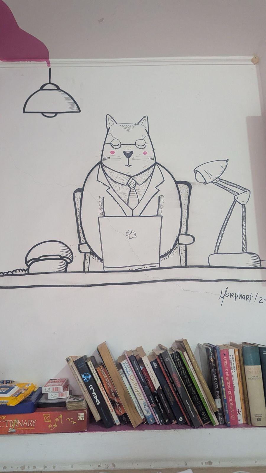 Wall mural of a cat working at a desk with a laptop, with bookshelves below, at Café Fundación in Medellin