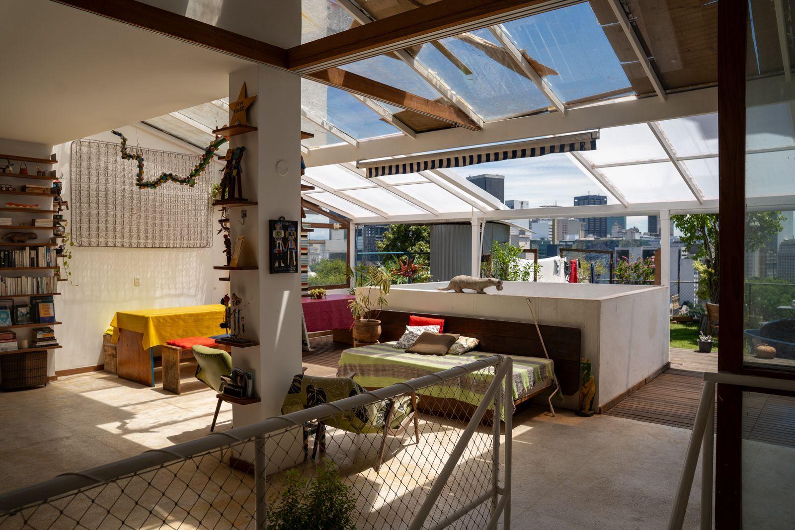 Open rooftop terrace with glass ceiling, colorful furniture, bookshelves, and city skyline views — artsy and homey atmosphere