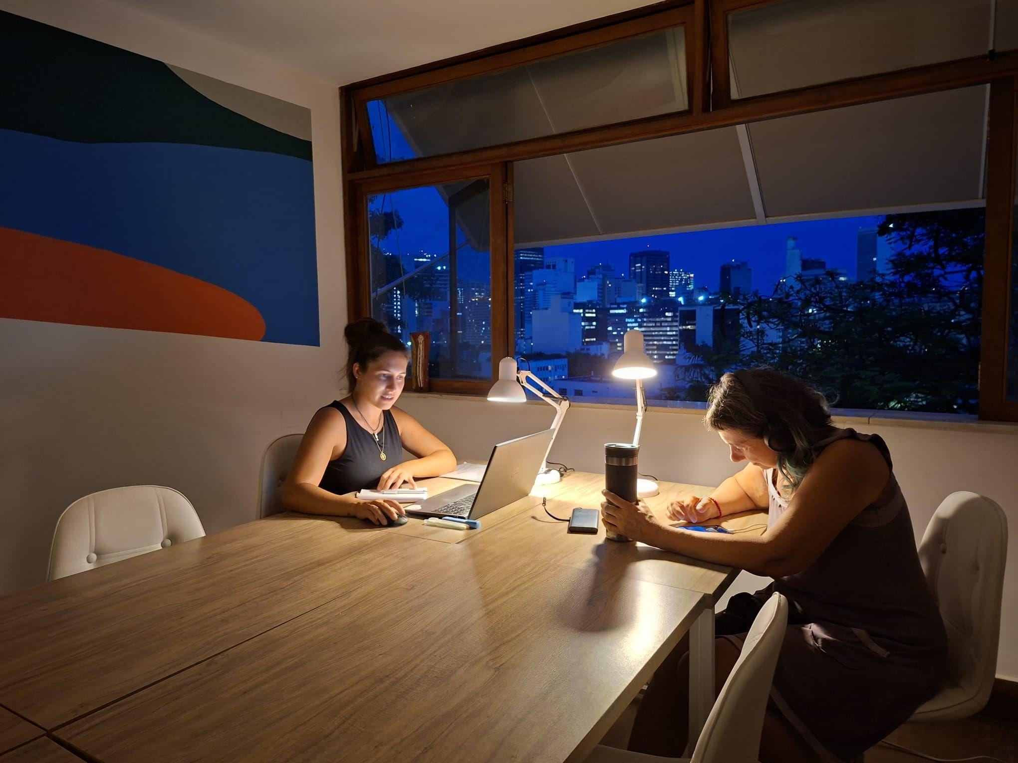 Two people working on laptops at the coworking table at night, with city lights visible through the large windows