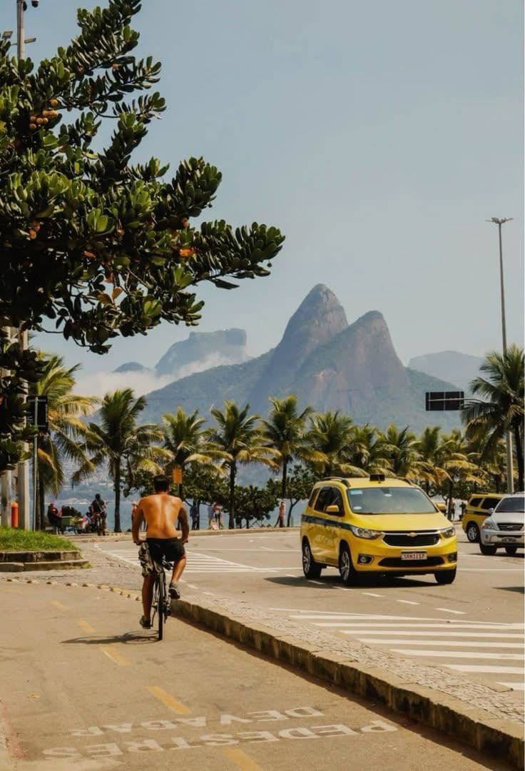 A cyclist and taxi on a Rio de Janeiro street with Morro Dois Irmãos mountain in the background, palm trees lining the road