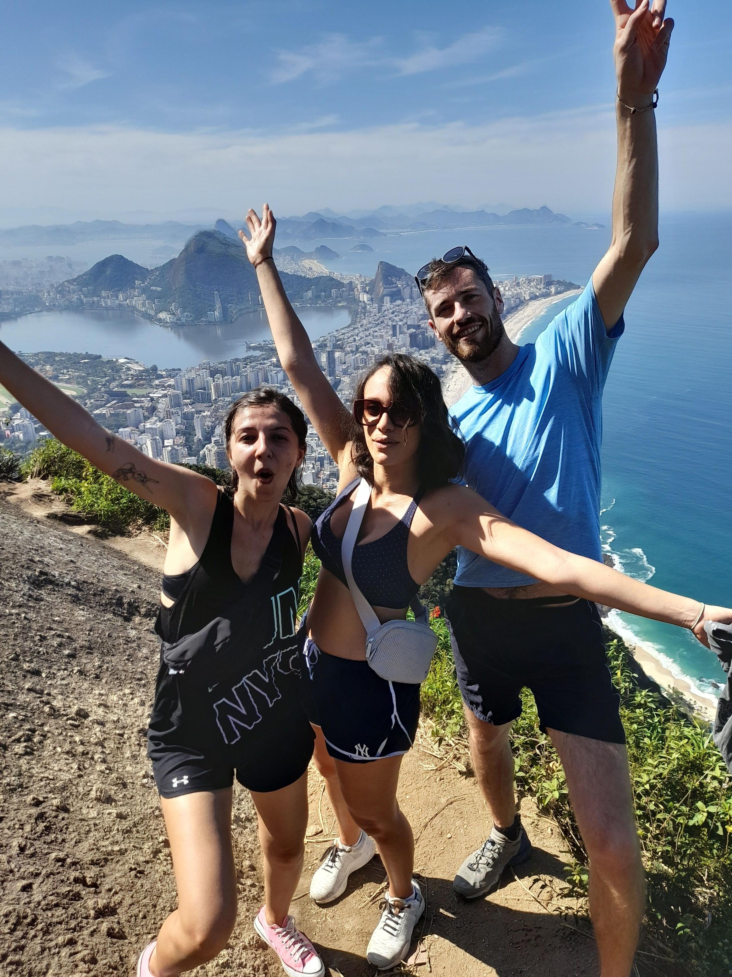 Three people celebrating at the top of Morro Dois Irmãos with panoramic views of Rio de Janeiro behind them