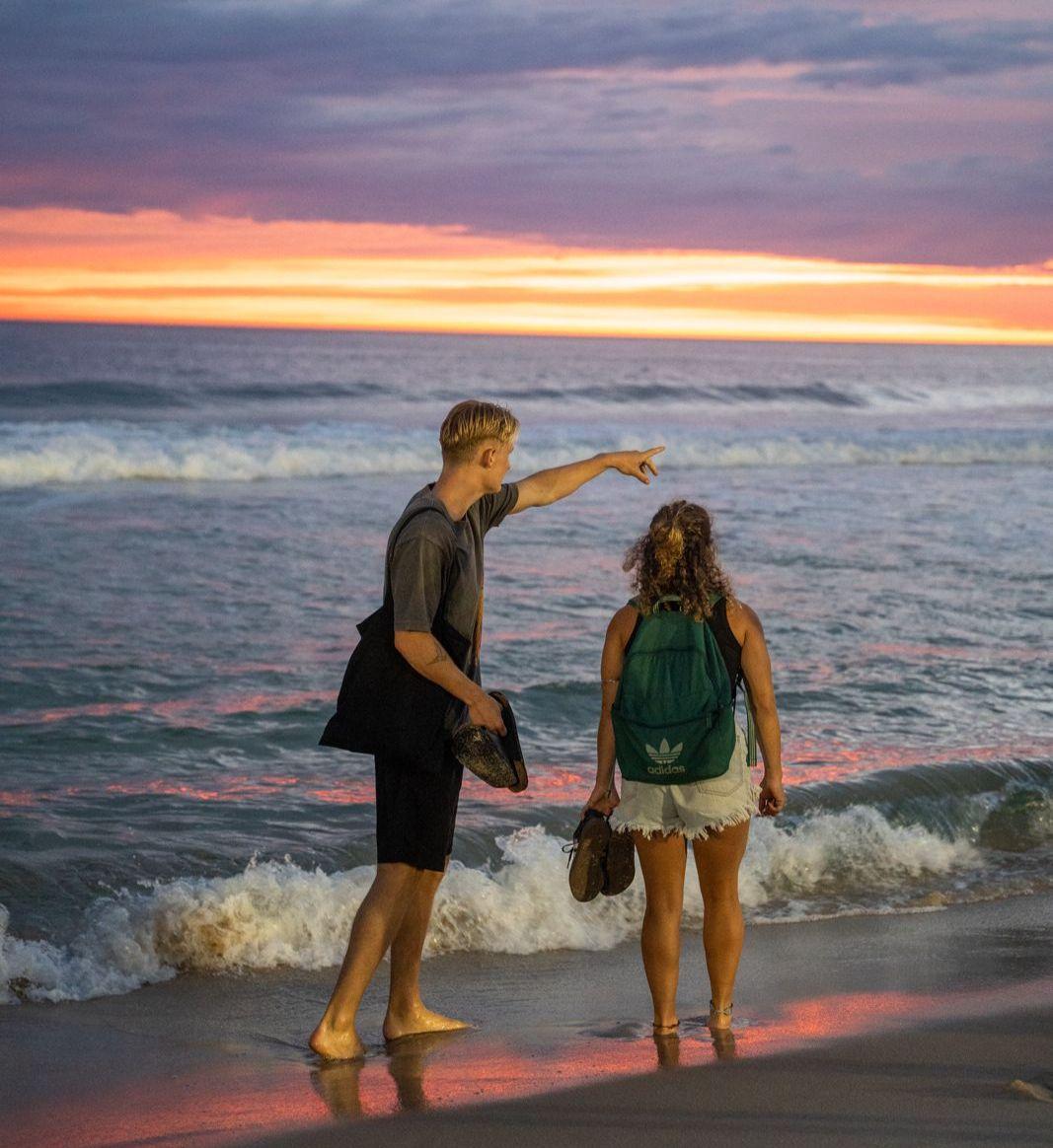 Two people standing at the shoreline watching a vivid sunset on a Rio de Janeiro beach