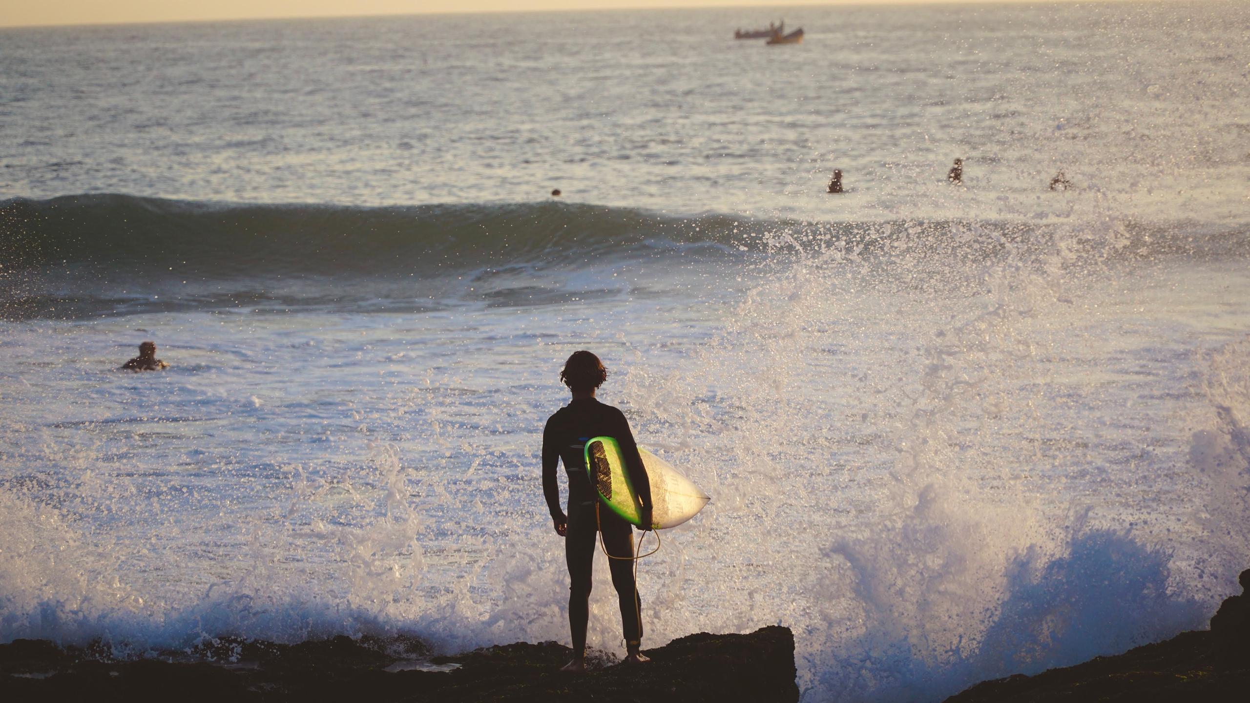 Surfer standing on rocks with surfboard watching the waves at sunset near Taghazout