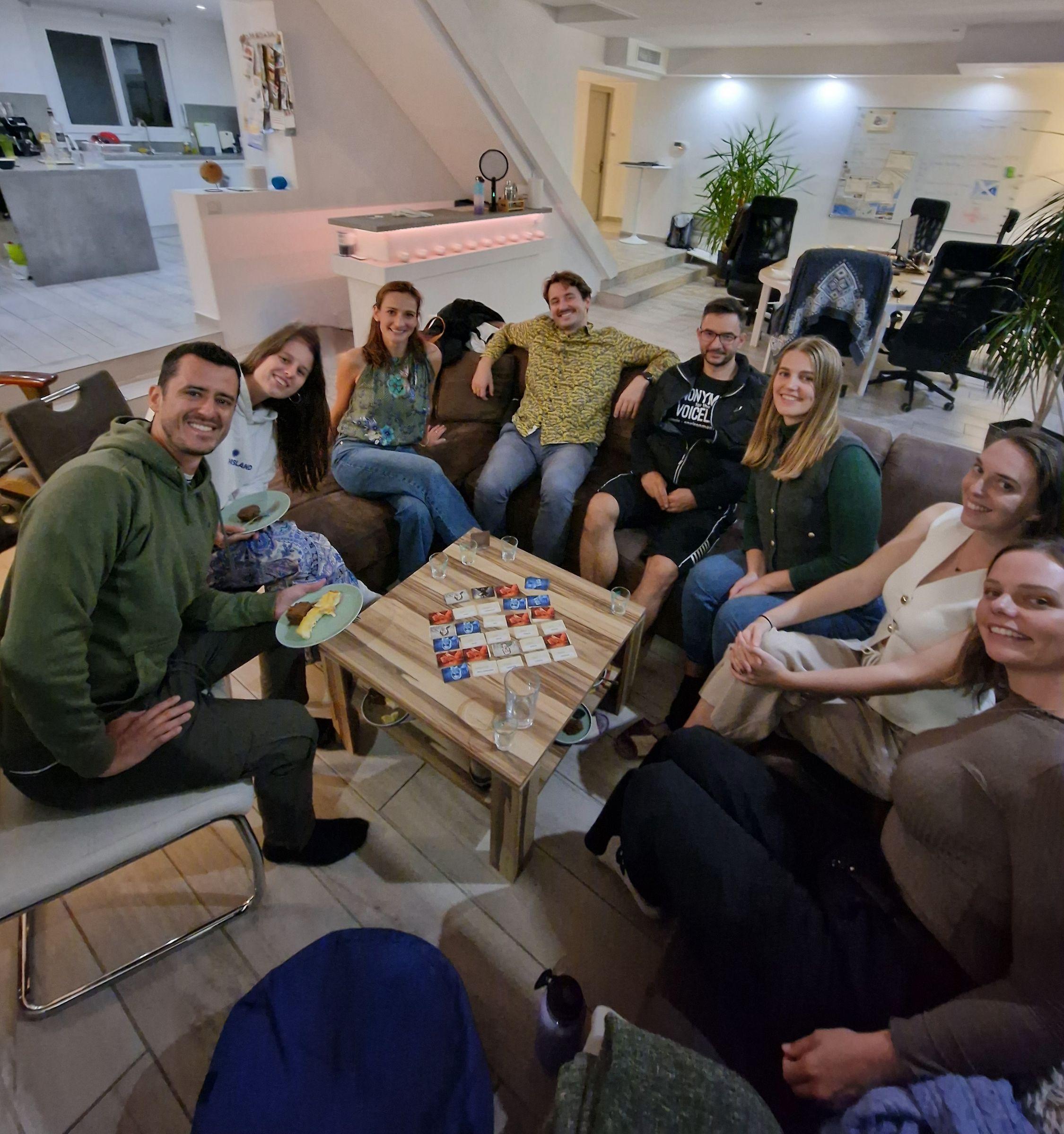 Group of coliving residents playing board games in the apartment living room, with kitchen and coworking area visible in the background