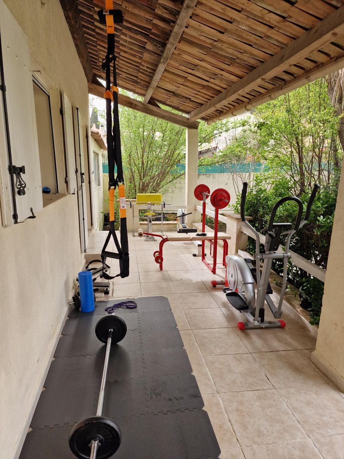 Outdoor fitness corner with weights, barbells, TRX straps, elliptical machine, and other gym equipment under a covered area