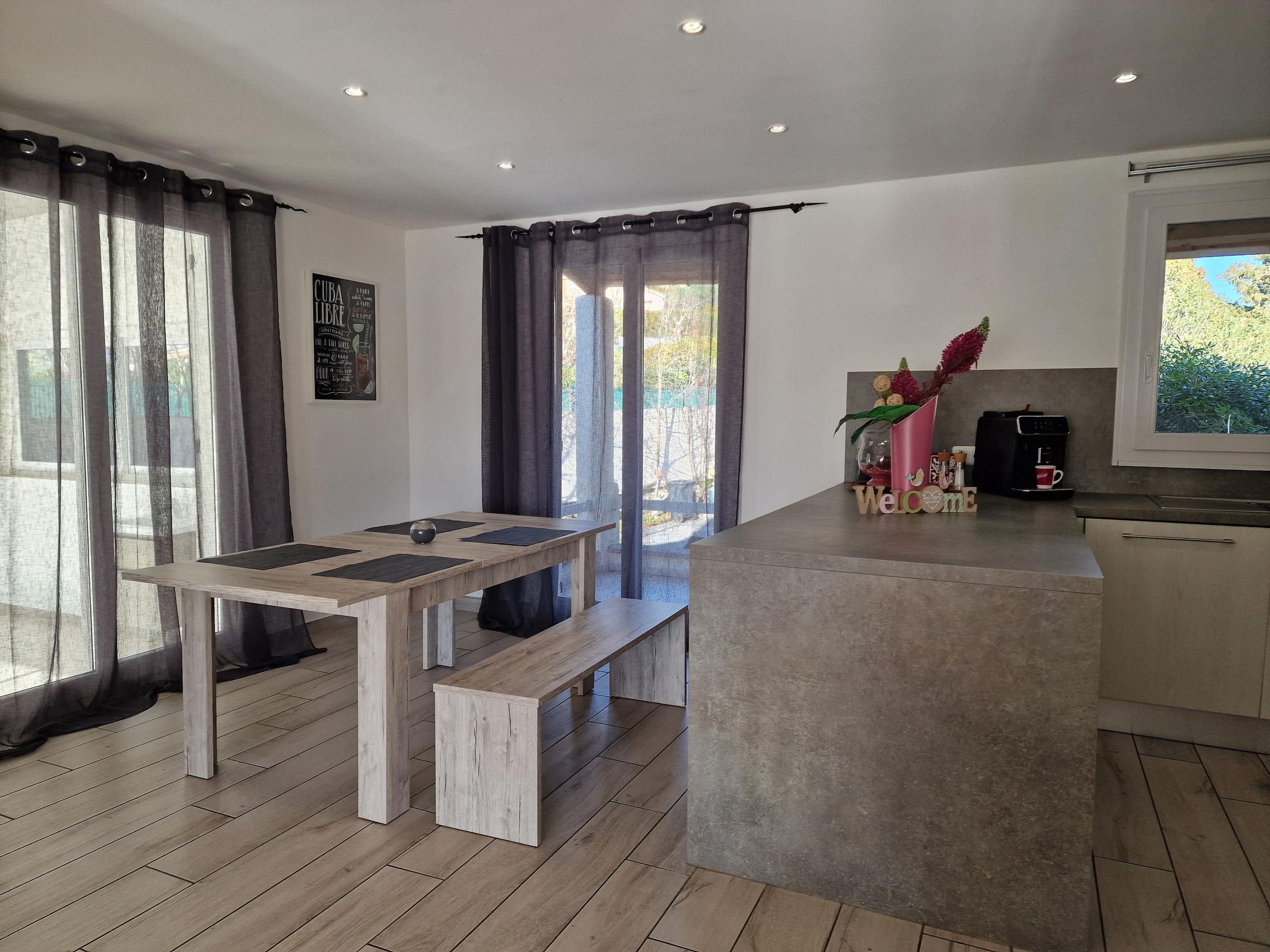 Dining area with a wooden bench table and kitchen island visible, with sliding doors leading to the terrace