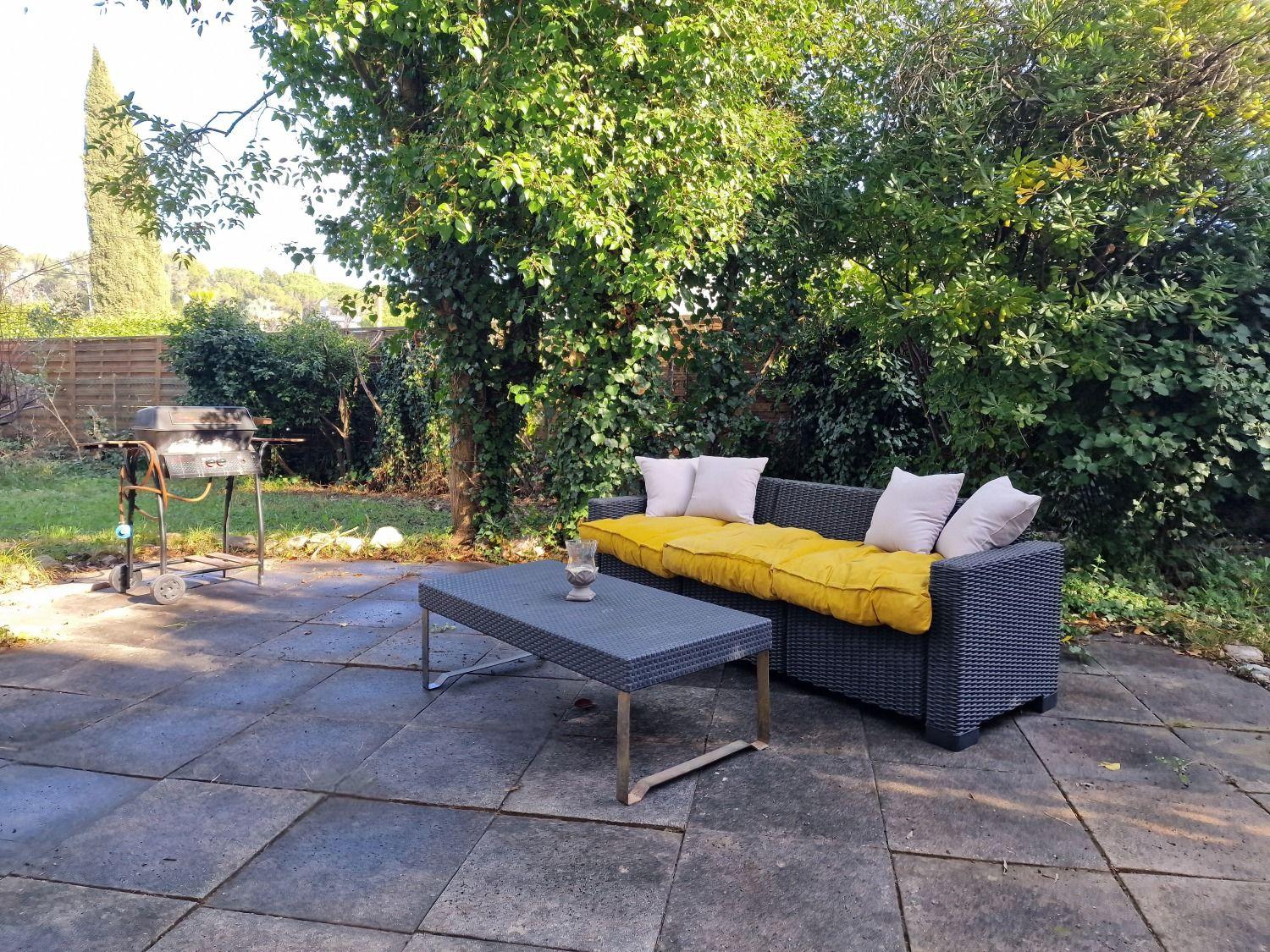 Garden lounge area with a sofa and coffee table surrounded by lush greenery and a BBQ grill