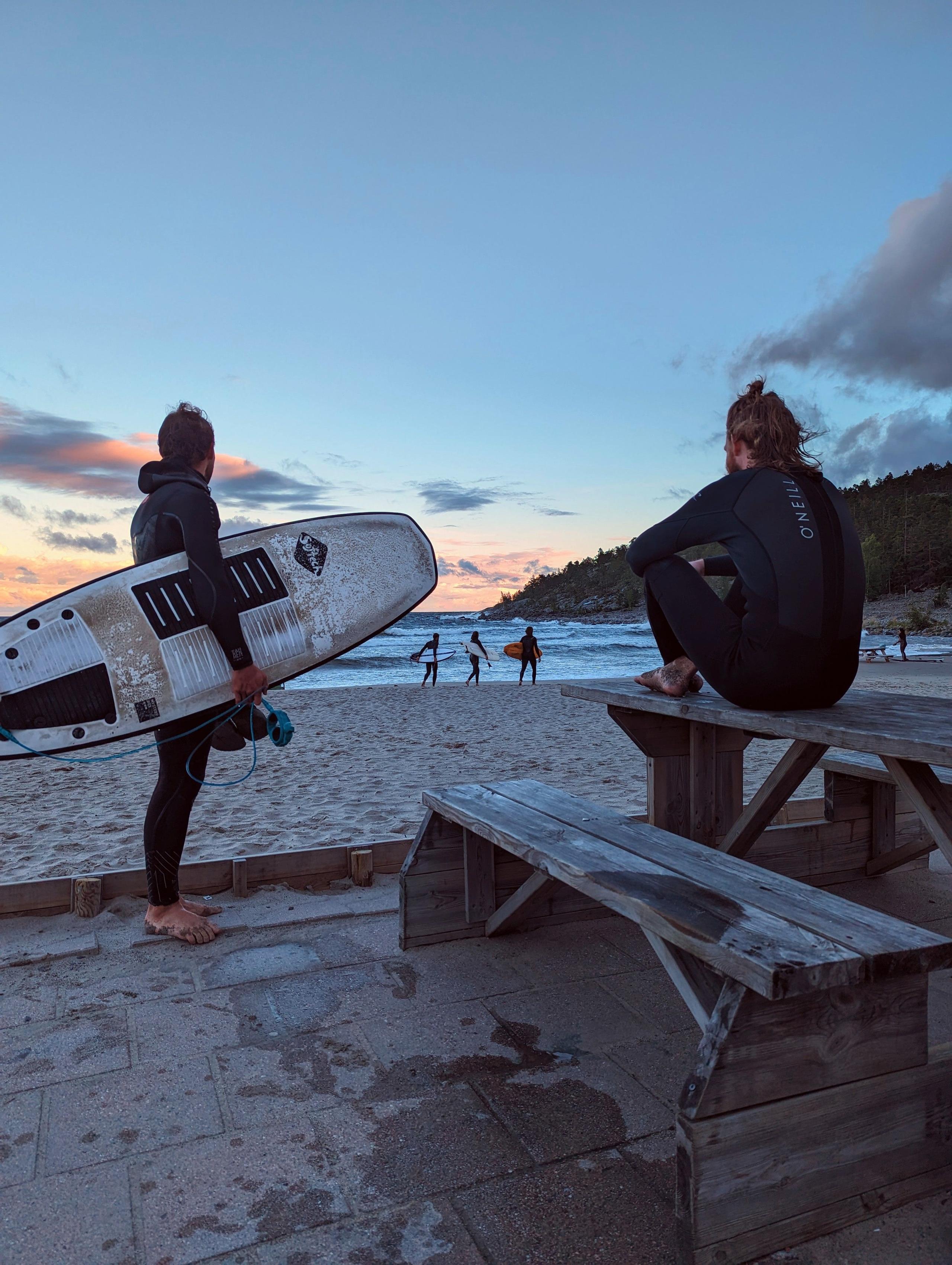Two surfers at the beach at sunset, one holding a surfboard, others surfing in the background