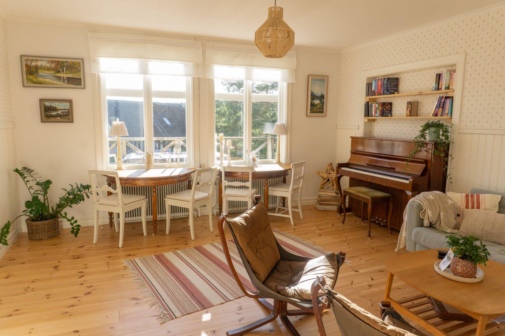 Bright shared living and coworking room with dining tables, piano, bookshelf, sofa and large windows