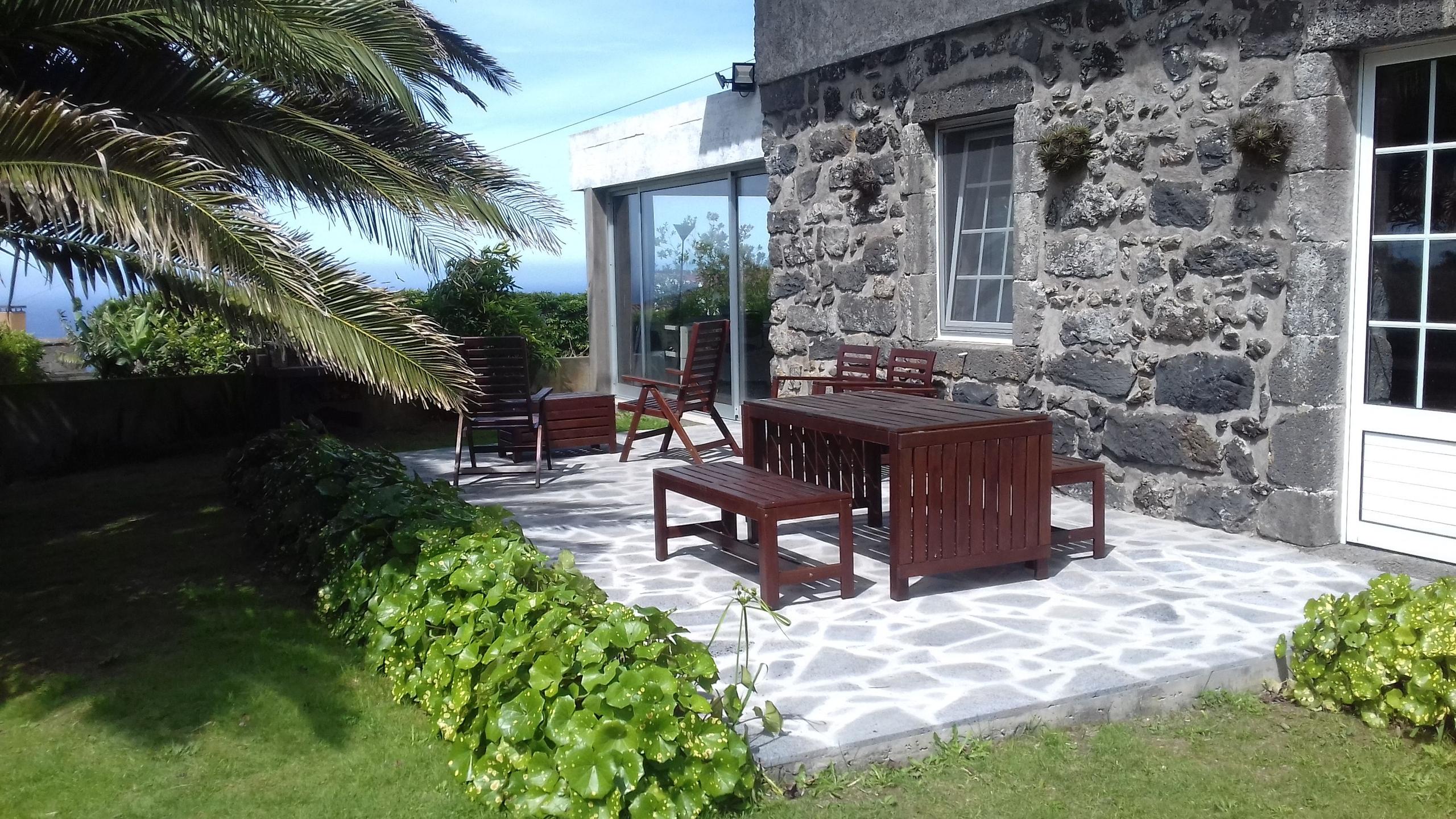 Stone-paved terrace with wooden outdoor furniture adjacent to the traditional Azorian stone house, with ocean view in the background