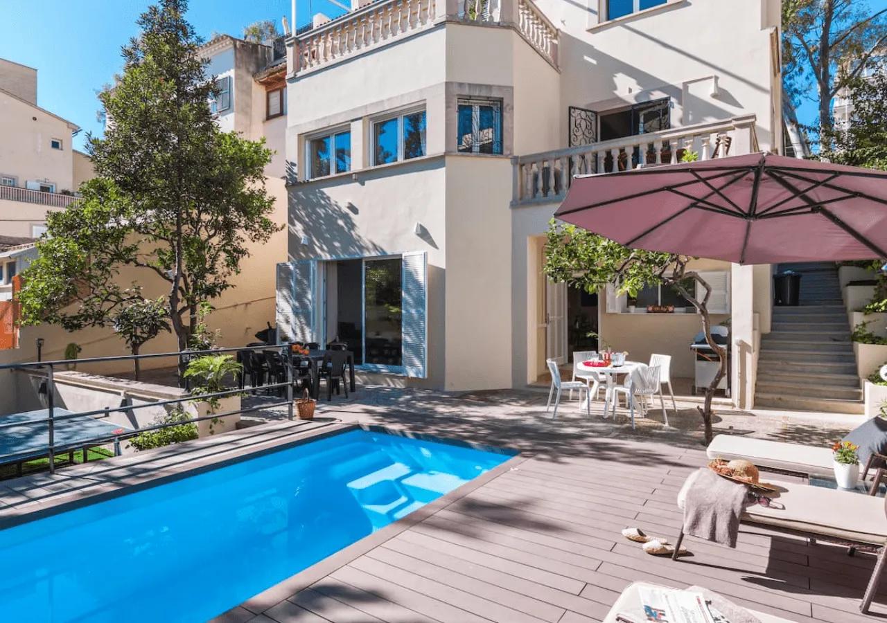 Daytime view of Villa Armadans garden with swimming pool, wooden deck, sun loungers, outdoor dining area, and the villa facade