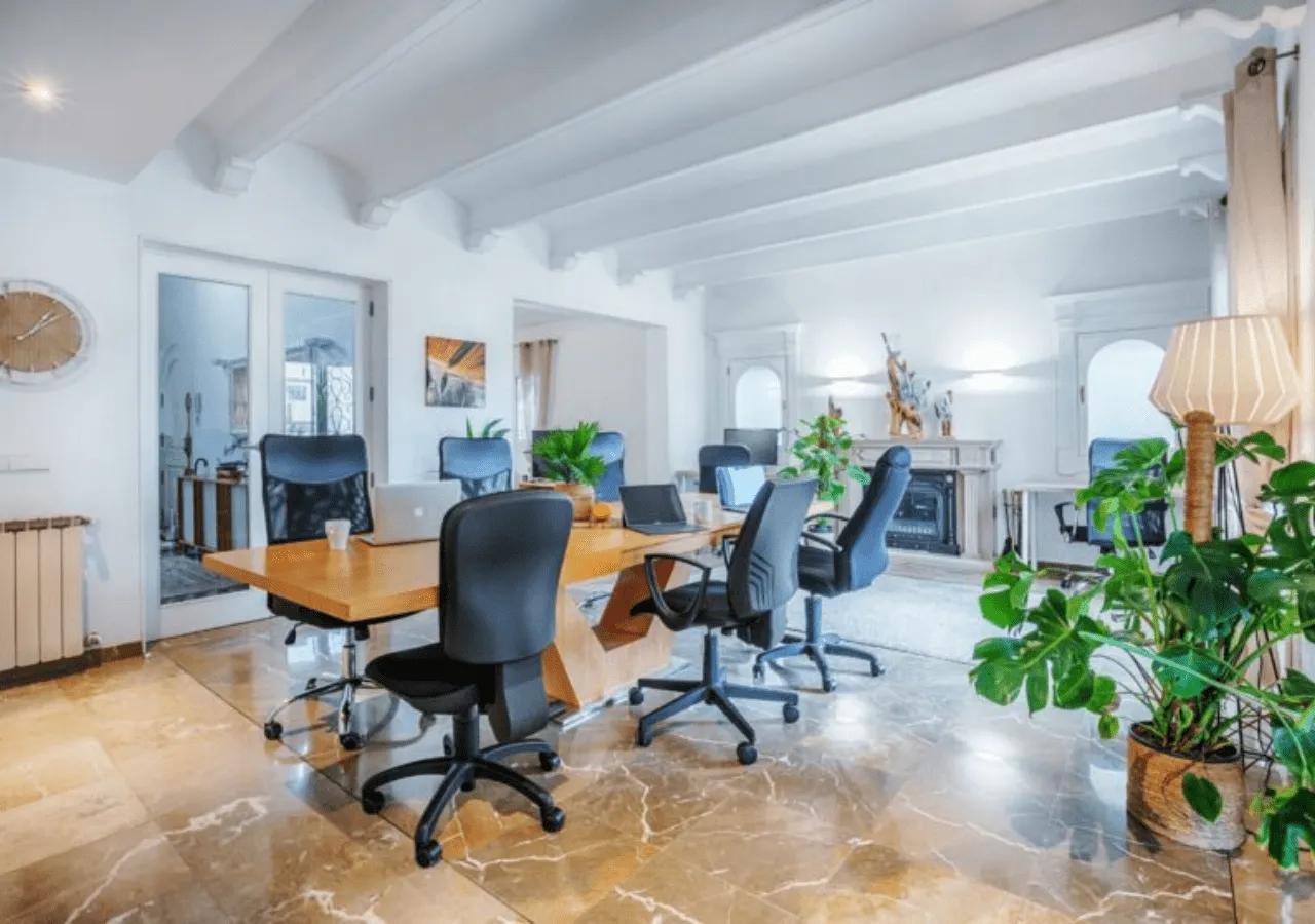 Coworking space with hot desks, ergonomic chairs, laptops, plants, and high ceilings with exposed beams, viewed from a different angle