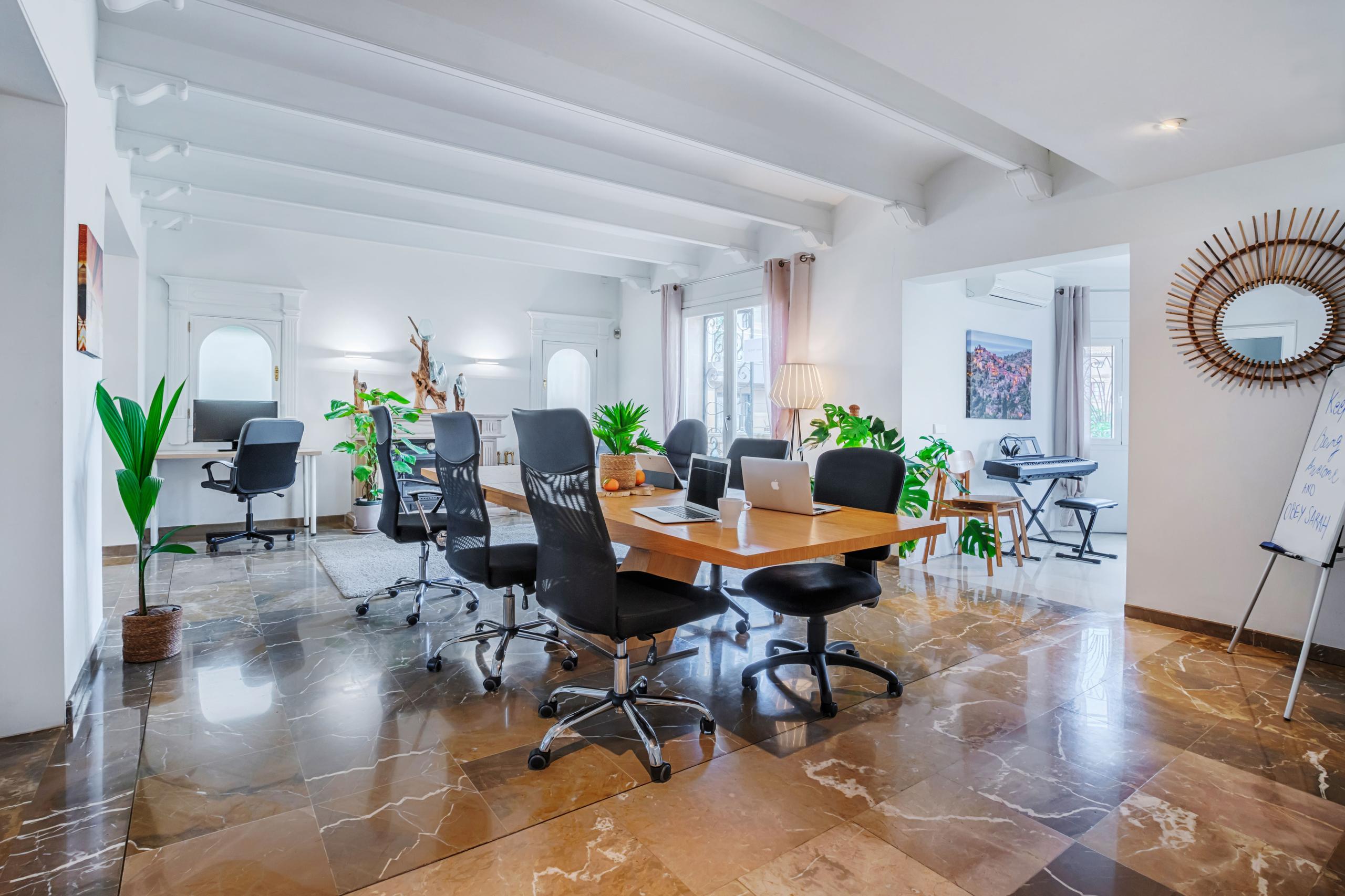 Spacious, light-filled coworking room with hot desks, ergonomic chairs, laptops, plants, and high ceilings with exposed beams
