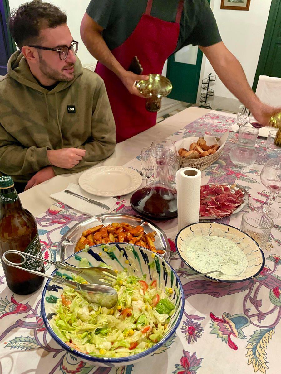 A resident at a dinner table with colourful ceramic bowls of salad, tapas and wine, served by someone in a red apron