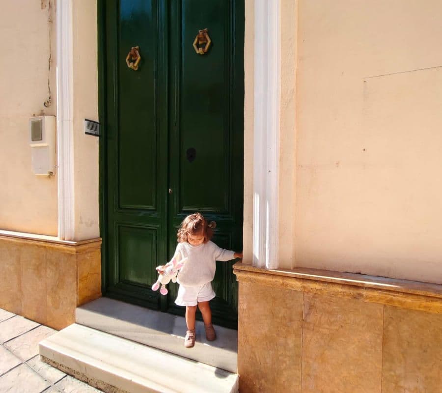 A child standing at the green double-door entrance of the Picassa coliving house, with warm stone facade