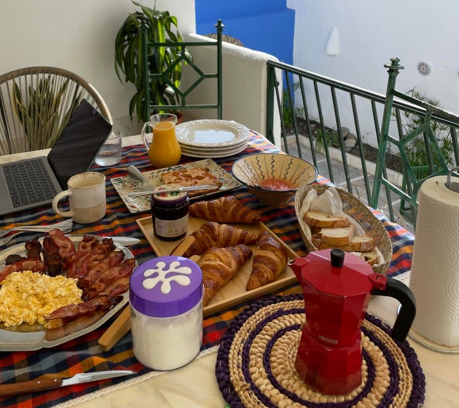 A full breakfast spread on the terrace table including croissants, scrambled eggs, bacon, orange juice, jam and a moka pot, with a laptop open