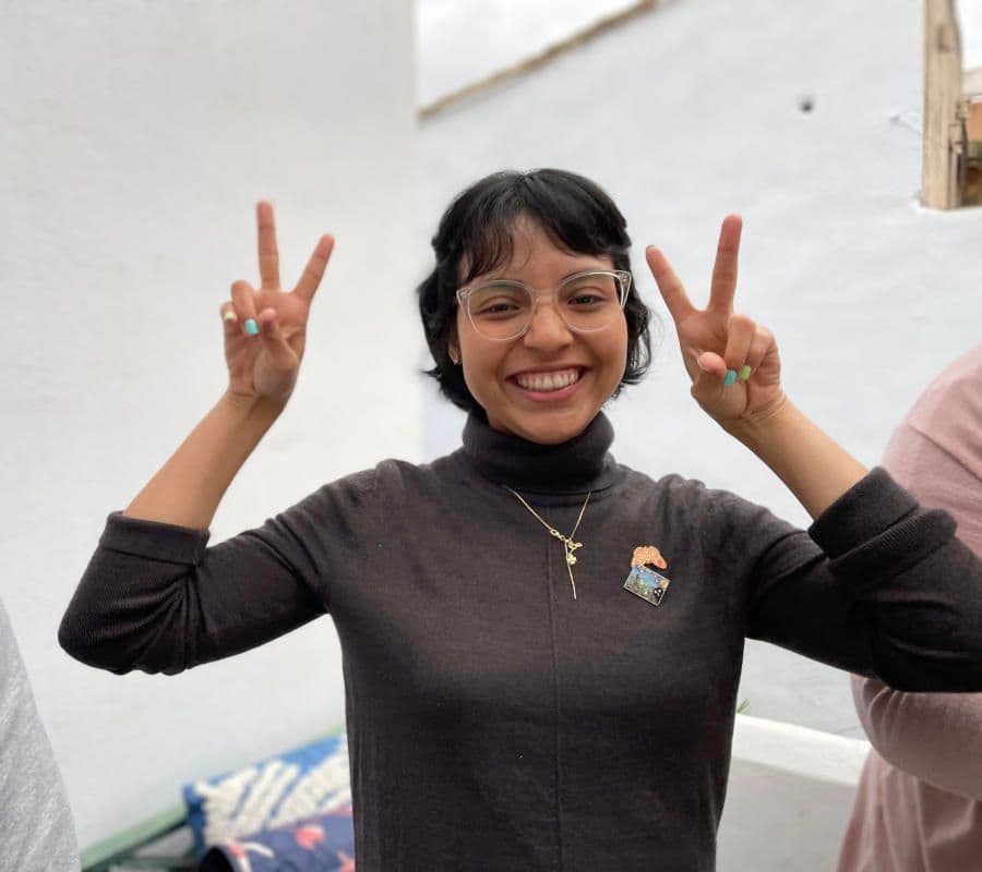A smiling woman with glasses making double peace signs, wearing a Purchena badge, at the Iberbibliotecas event