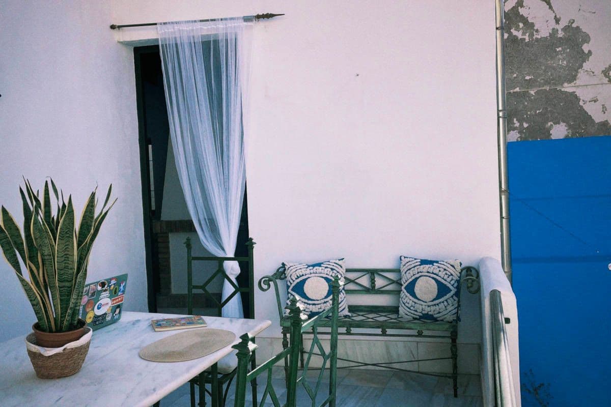 A small outdoor terrace with a marble table, iron bench with decorative cushions, a plant, and sheer curtains