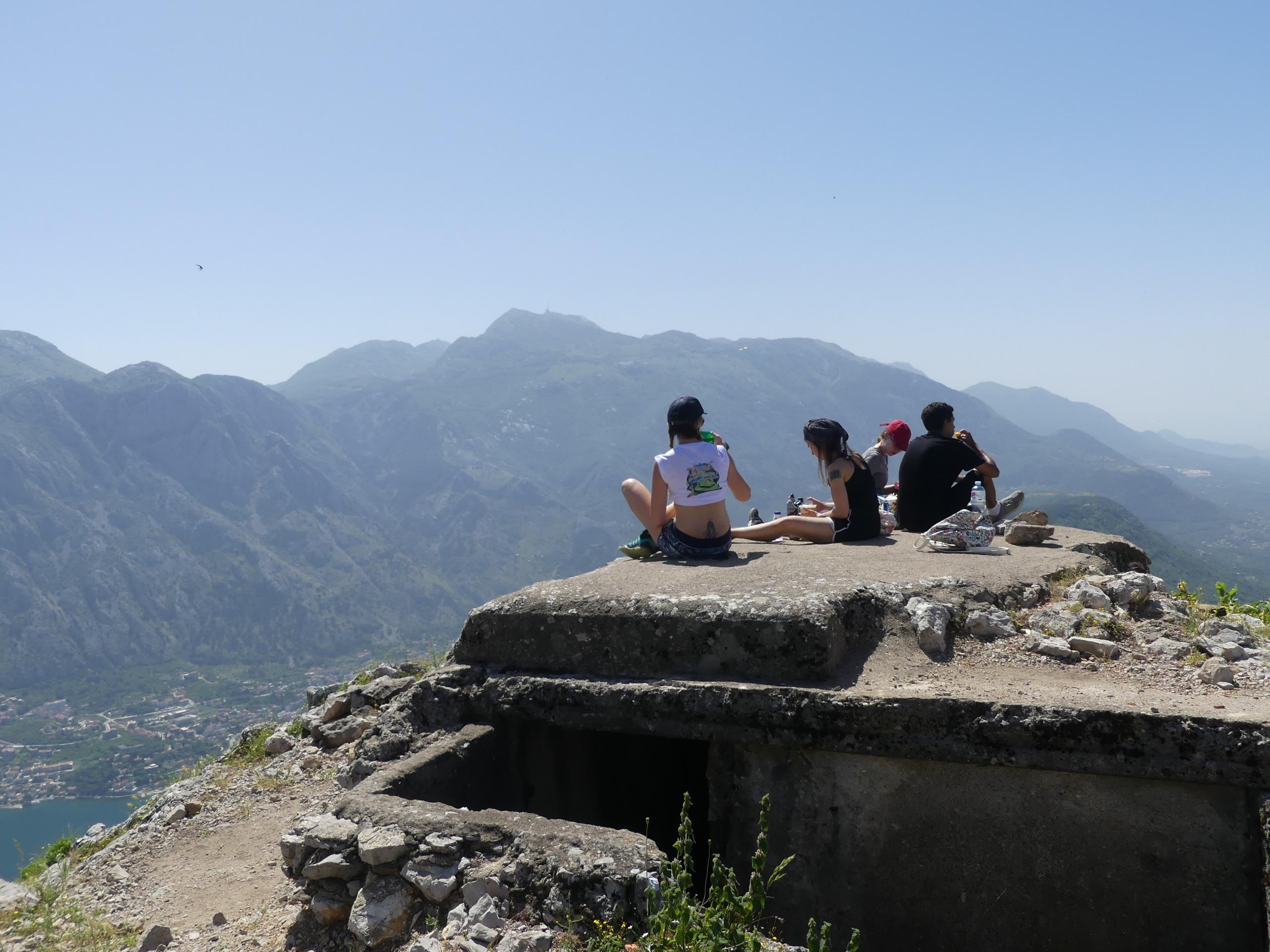 Group of people sitting on a rocky ledge at a mountain viewpoint overlooking Kotor Bay