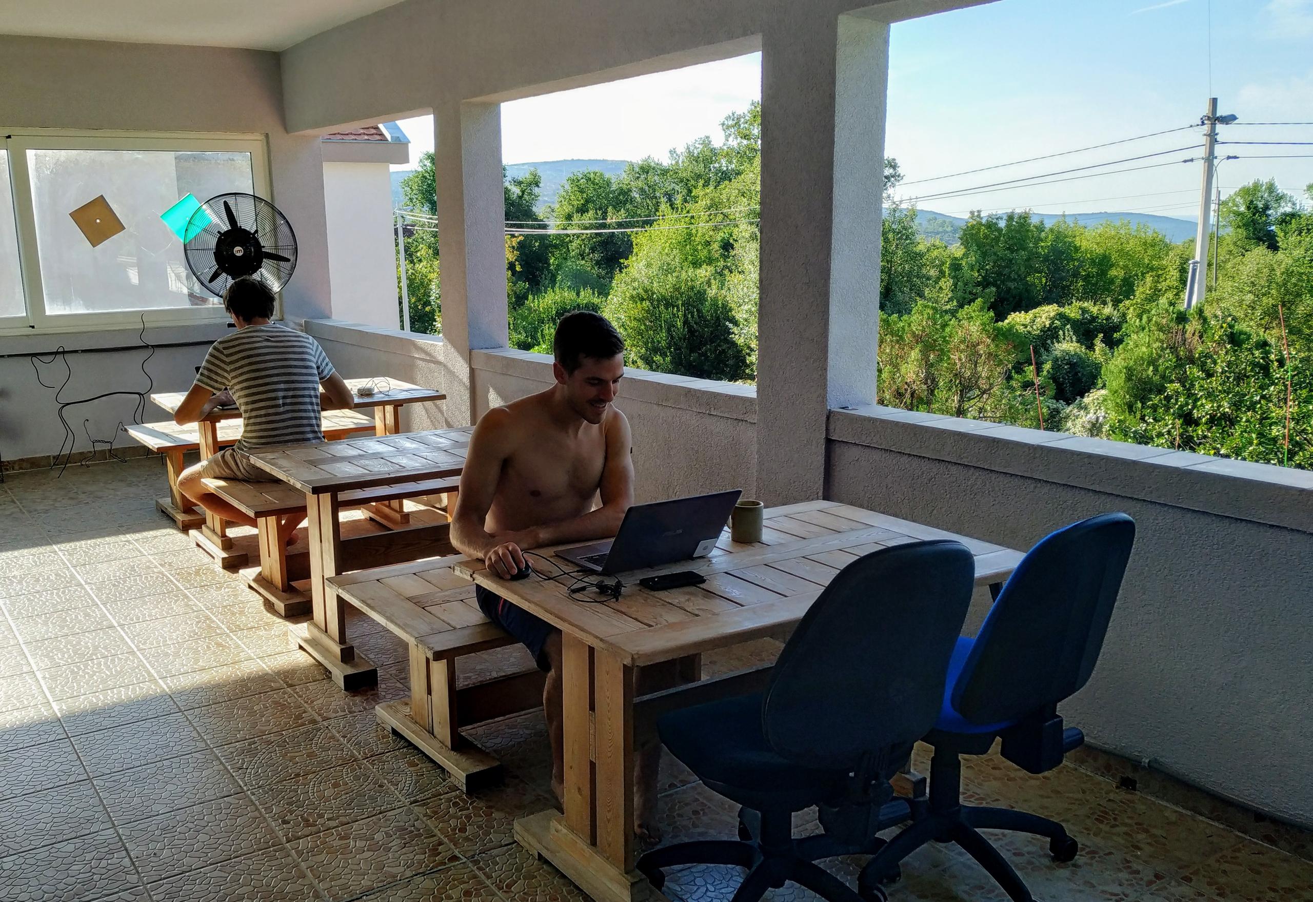 Covered outdoor terrace with wooden workstations and residents working on laptops, overlooking greenery