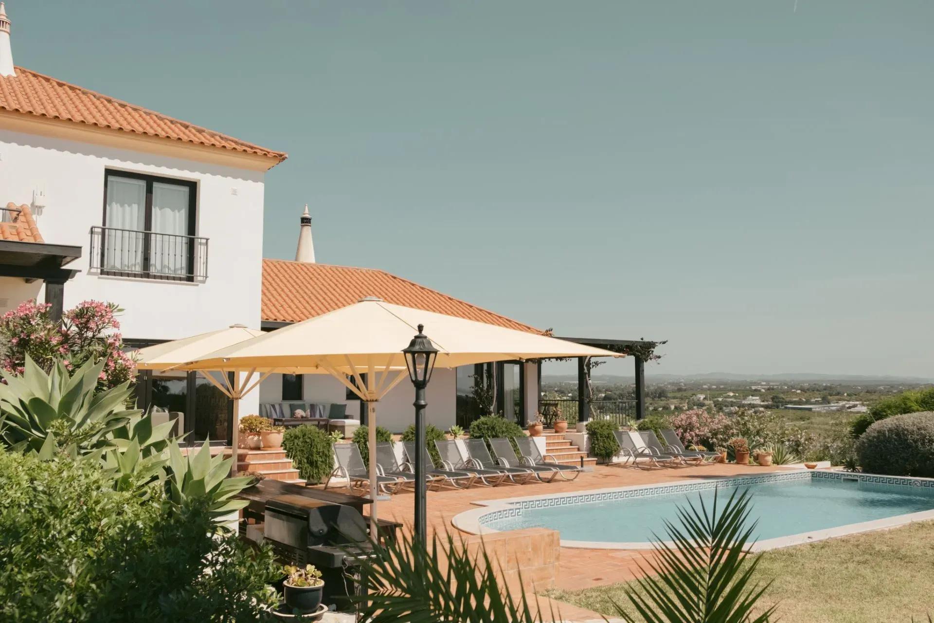 Mediterranean-style white villa at POMAR na praia with terracotta roof, shady umbrellas, sun-loungers and a turquoise swimming pool overlooking the Algarve countryside.