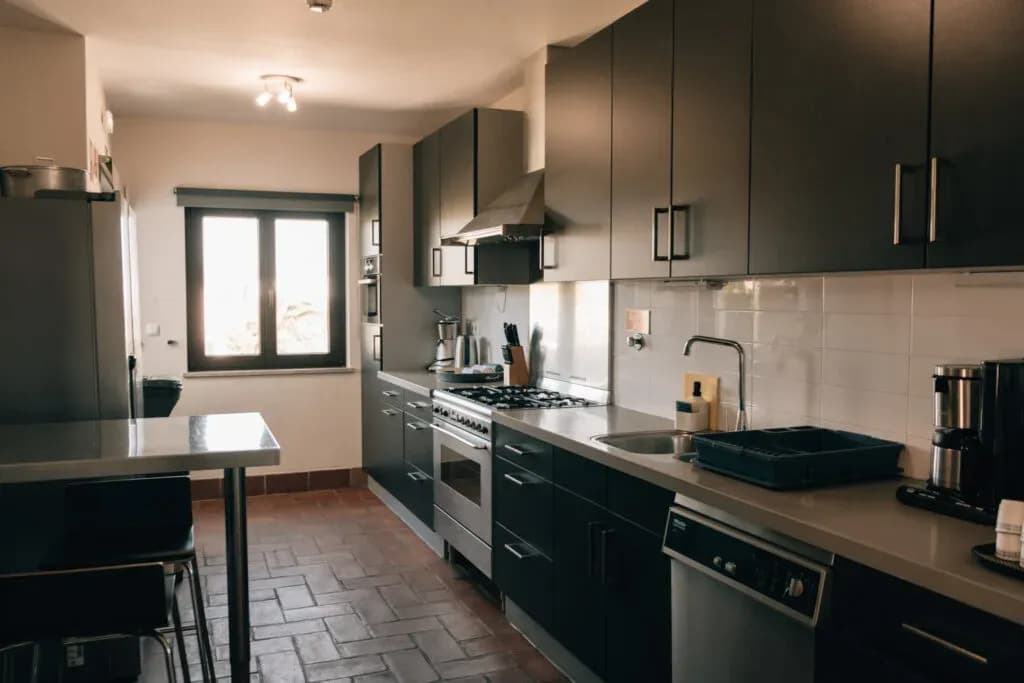 Modern fully equipped shared kitchen at POMAR na praia coliving, featuring dark cabinets, stainless steel appliances, and natural light from a large window.