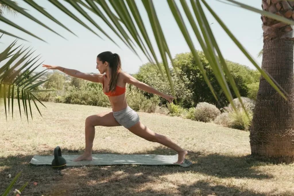 Person practicing yoga in a warrior pose on a mat outdoors at POMAR Coliving, surrounded by palm trees and nature