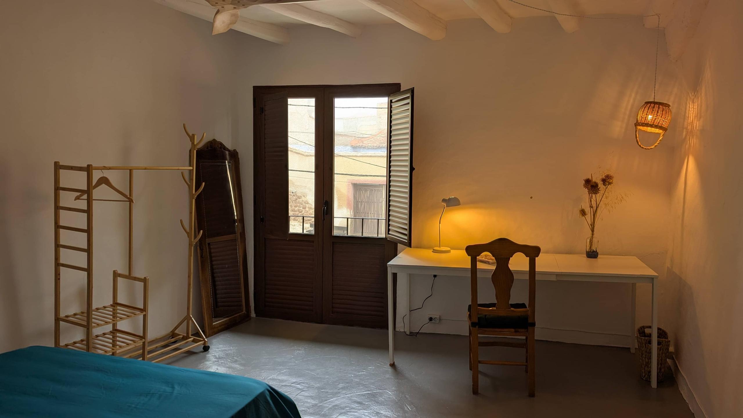The Graciela room showing a balcony window, study desk, lamp, chair, open books, dried flowers, a mirror on the floor and connectivity cube