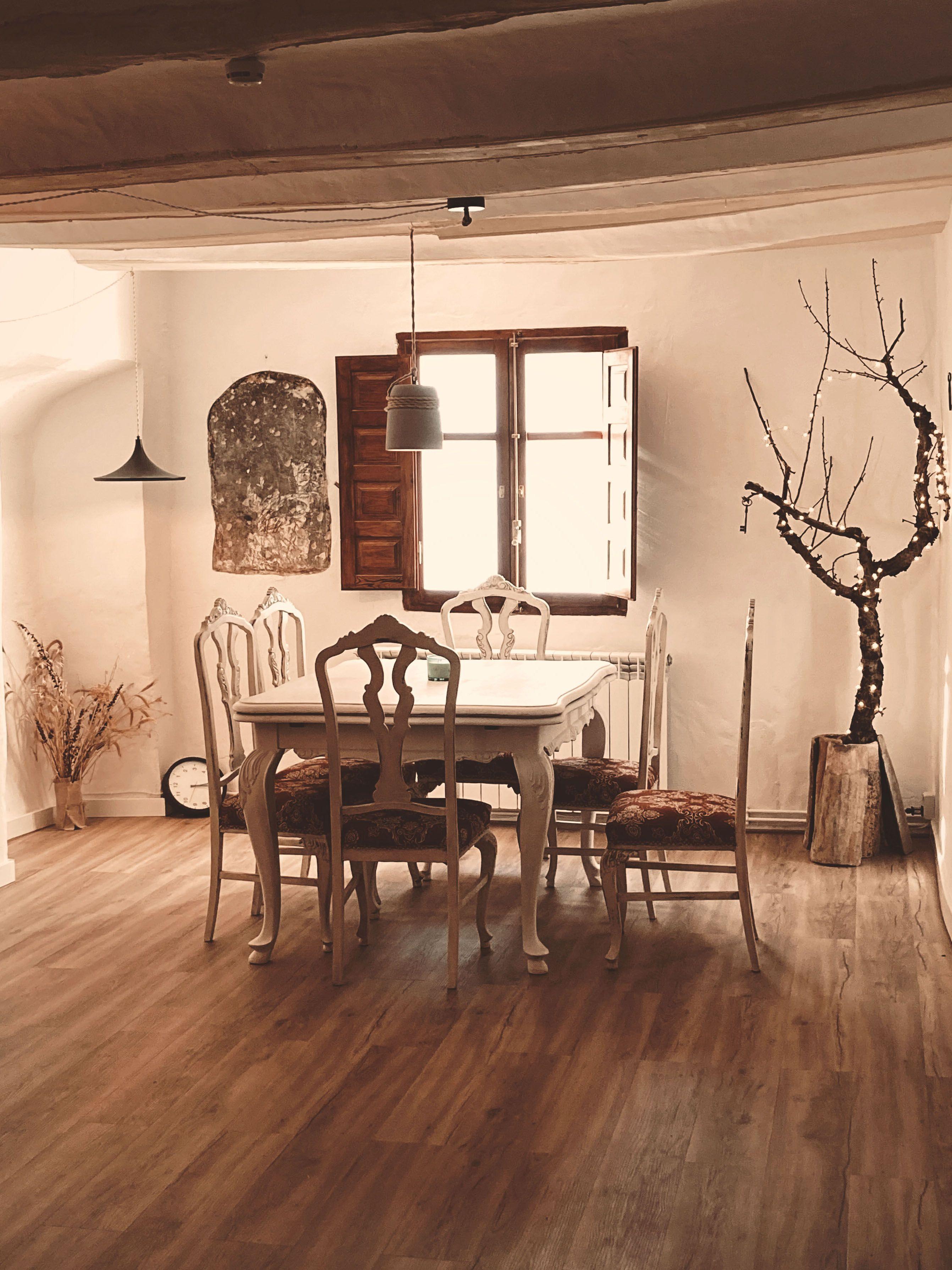 Work desk with chairs, window in the background, a dried tree on one side and a vase with dried flowers on the floor