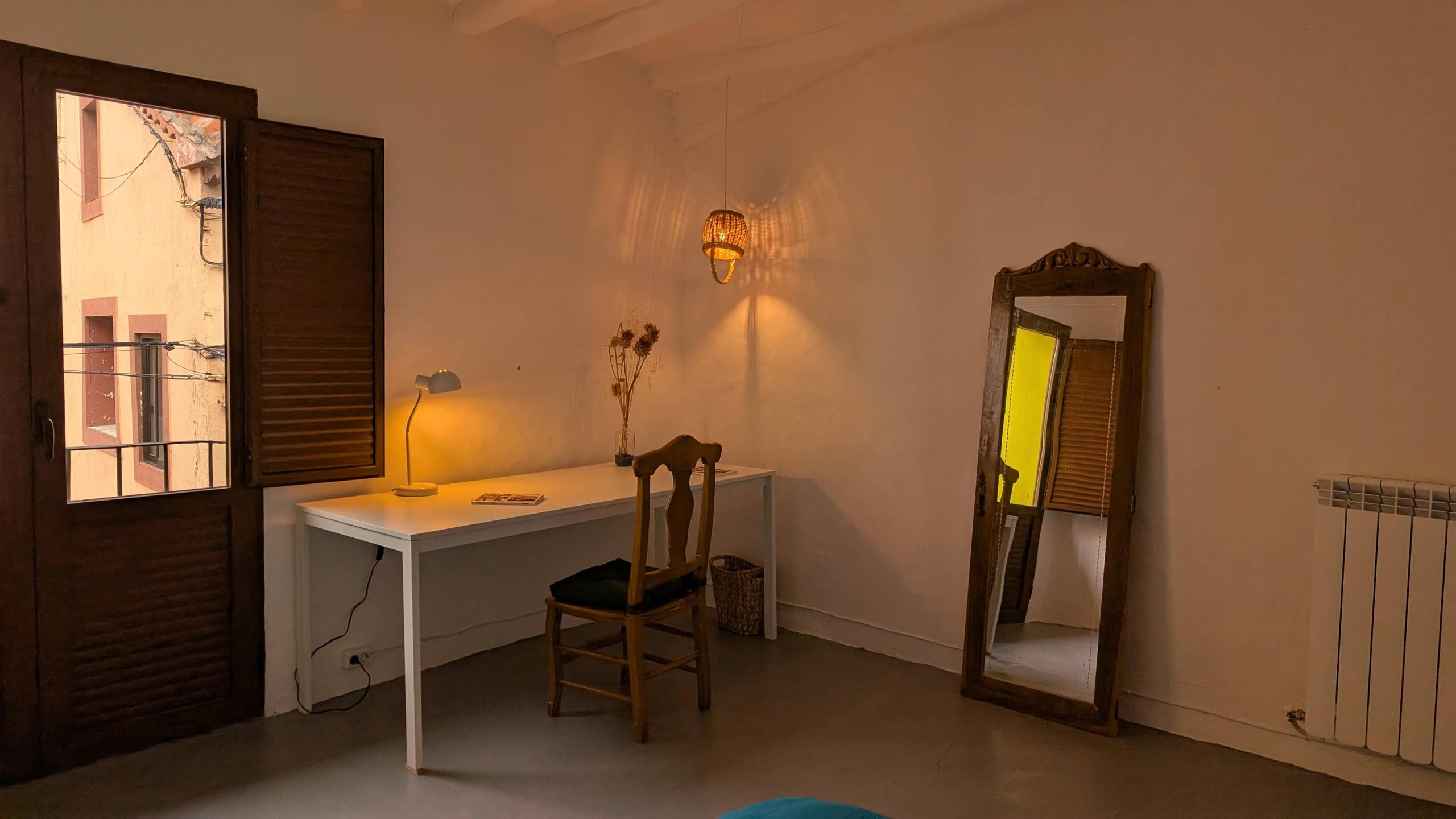 The Graciela room showing a balcony window, study desk, lamp, chair, open books, dried flowers, a mirror on the floor and connectivity cube