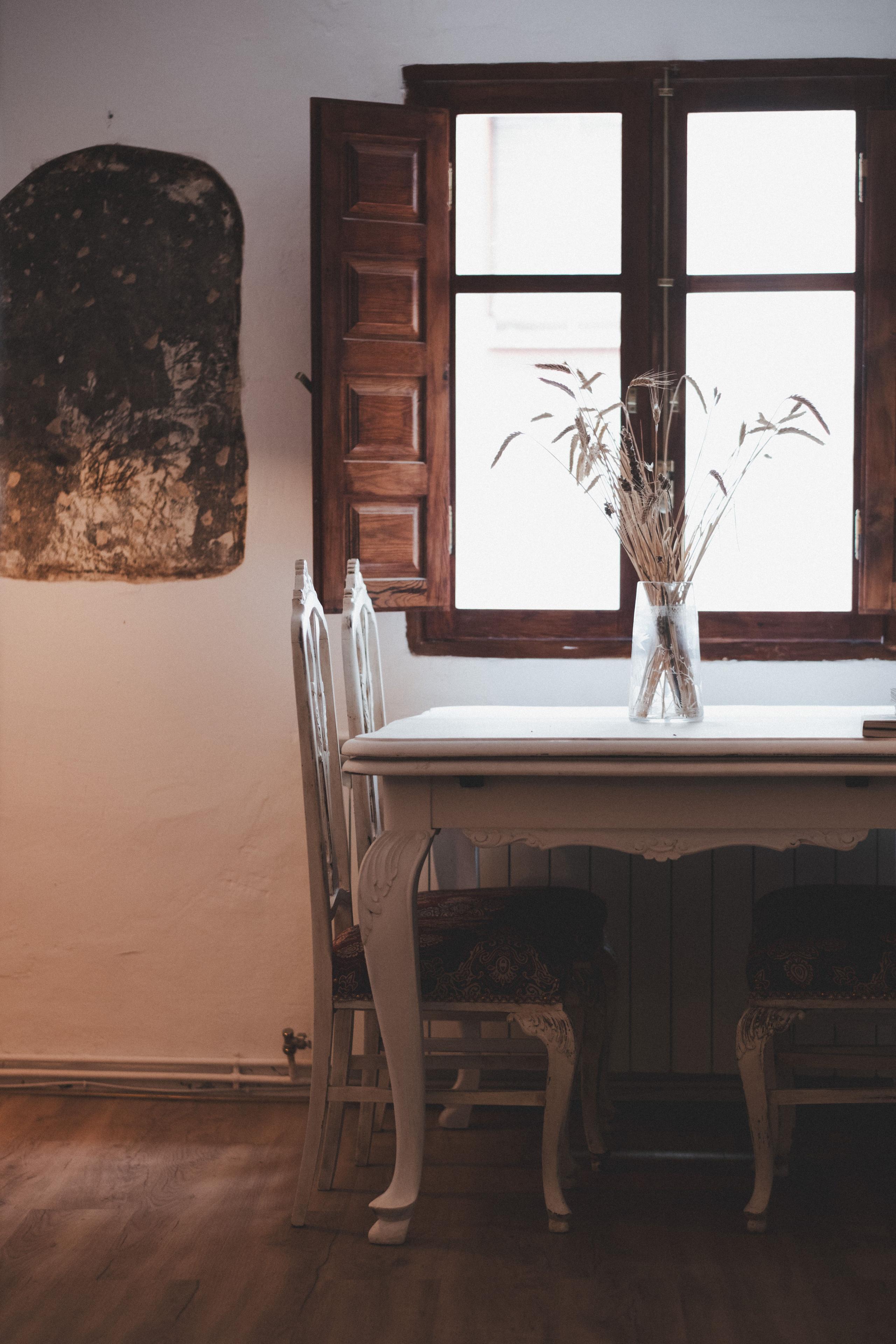 White work table with a vase of dried flowers in front of a window, surrounded by chairs, at Posada de la Luz