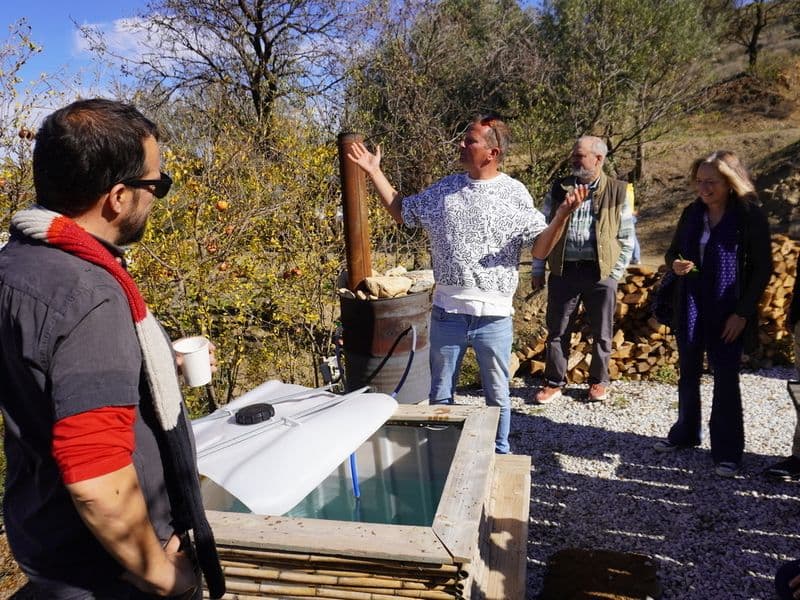 Group of people gathered around the outdoor hot tub, with one person explaining something, surrounded by trees and firewood