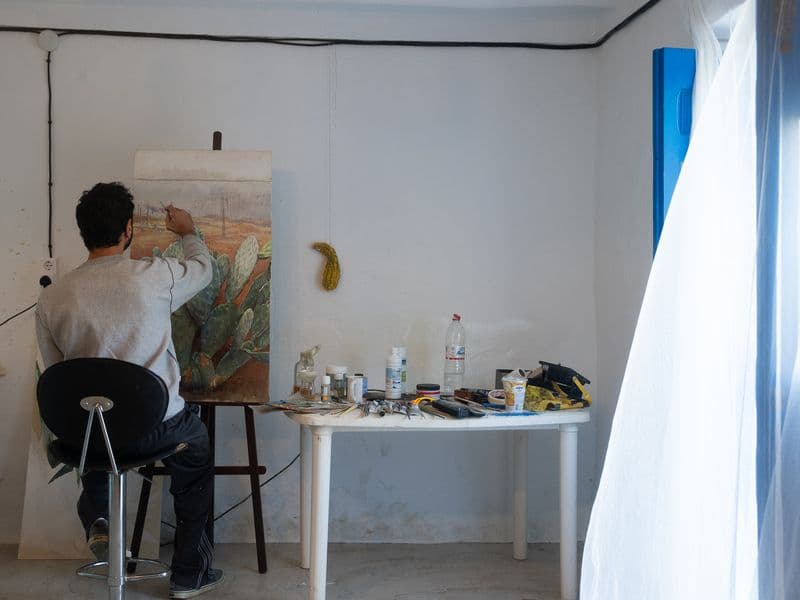 Artist sitting at an easel painting a cactus landscape in the studio, with art supplies on a table beside him