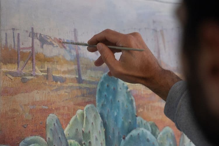 Close-up of an artist's hand holding a brush, painting a cactus landscape on canvas