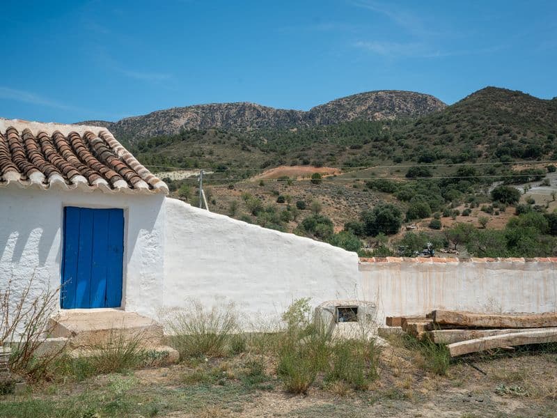 White-washed Andalusian building exterior with a blue door, terracotta roof tiles, and mountain backdrop