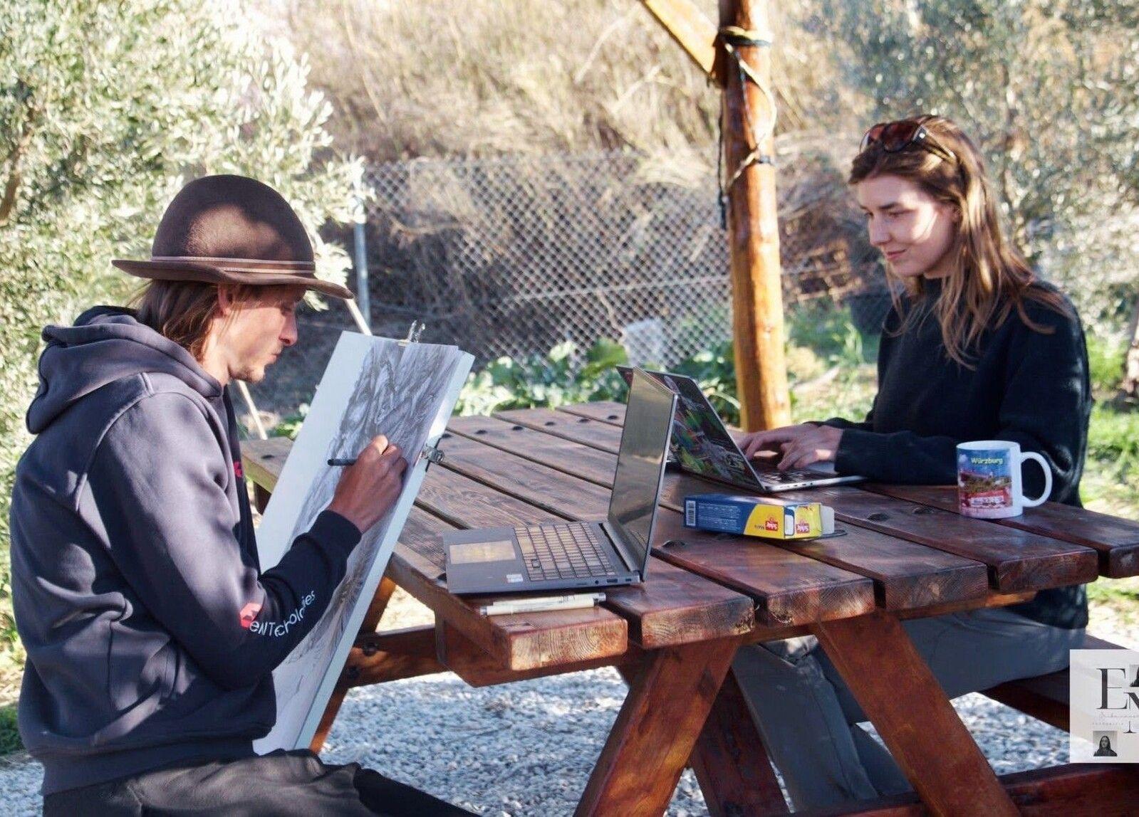 Two people working at an outdoor picnic table — one sketching in a notebook, the other on a laptop