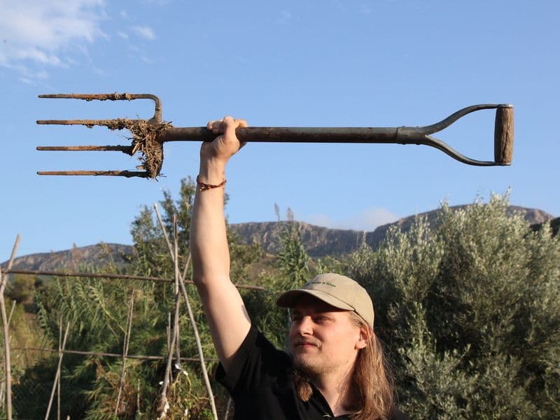 Person holding up a garden fork triumphantly in the permaculture garden with olive trees and mountains in the background