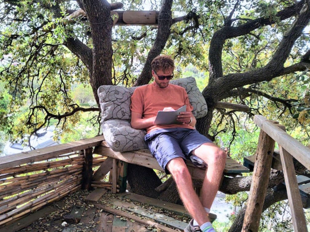 Man reading a book while seated on the tree platform installation, surrounded by tree branches