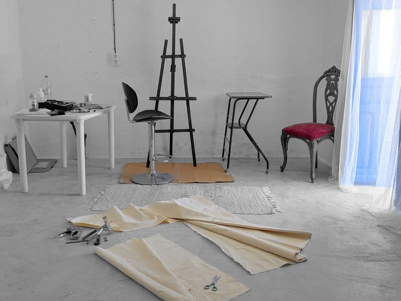 Empty artist studio with an easel, chairs, and canvas rolls on the floor, with blue curtains and natural light