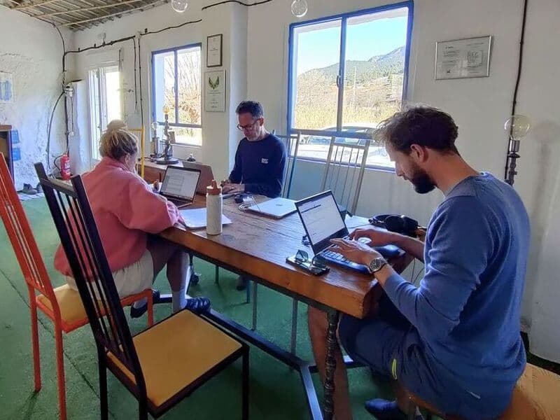 Three people working on laptops at a long wooden table in the gallery coworking space, with mountain views through large windows