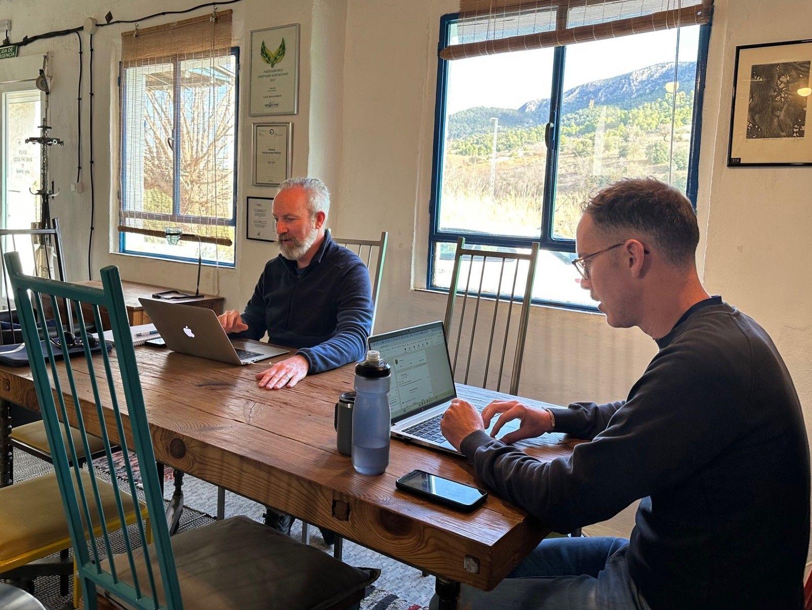 Two men working on laptops at a long wooden table inside the gallery coworking space, with mountain views through the windows
