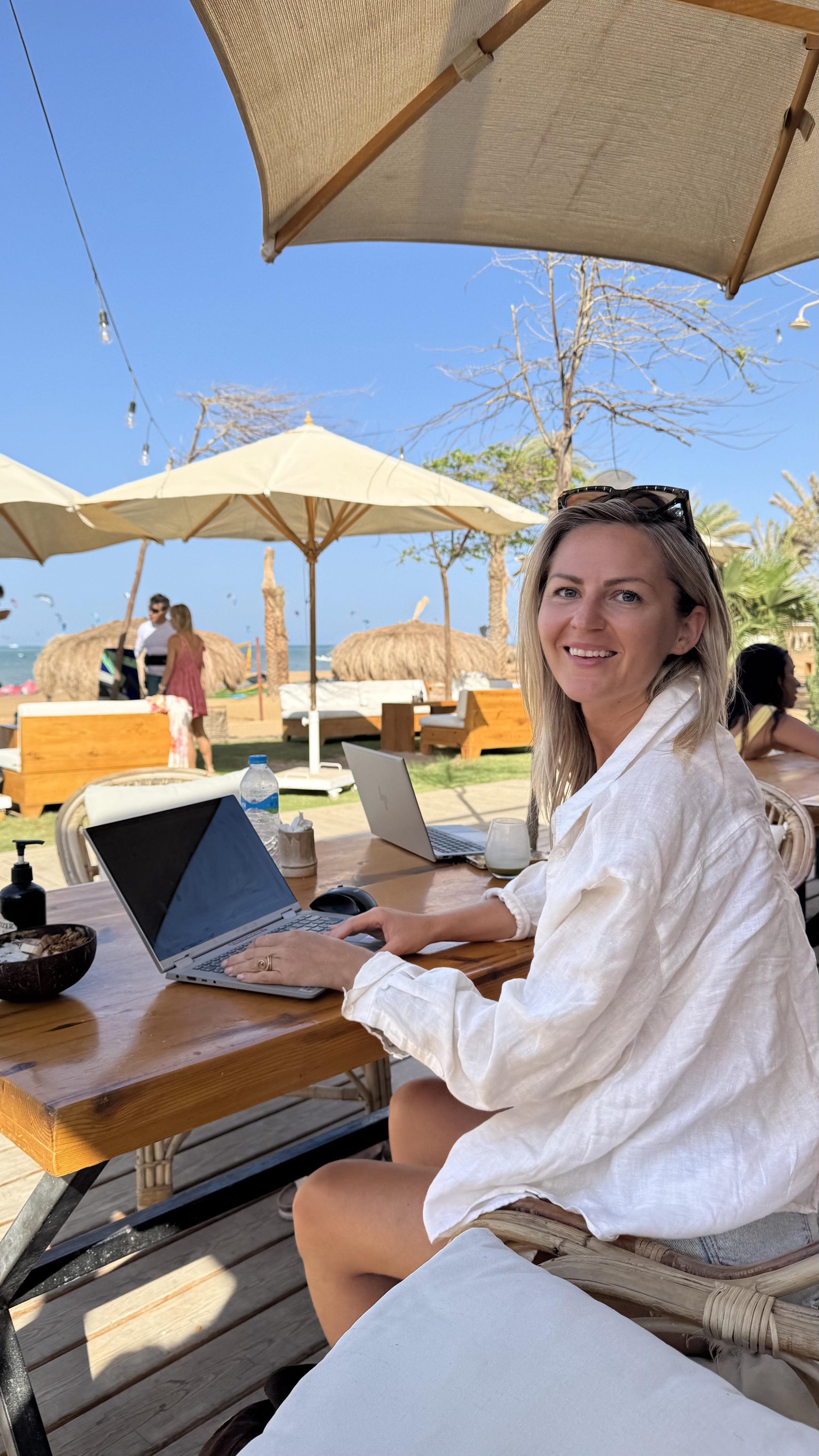 Person working on a laptop at a beach café with sea and palm trees in the background