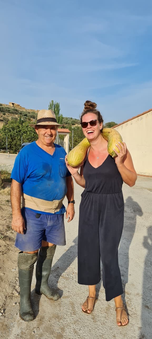 A digital nomad and a local grandpa are holding vegetables they picked from a local organic garden