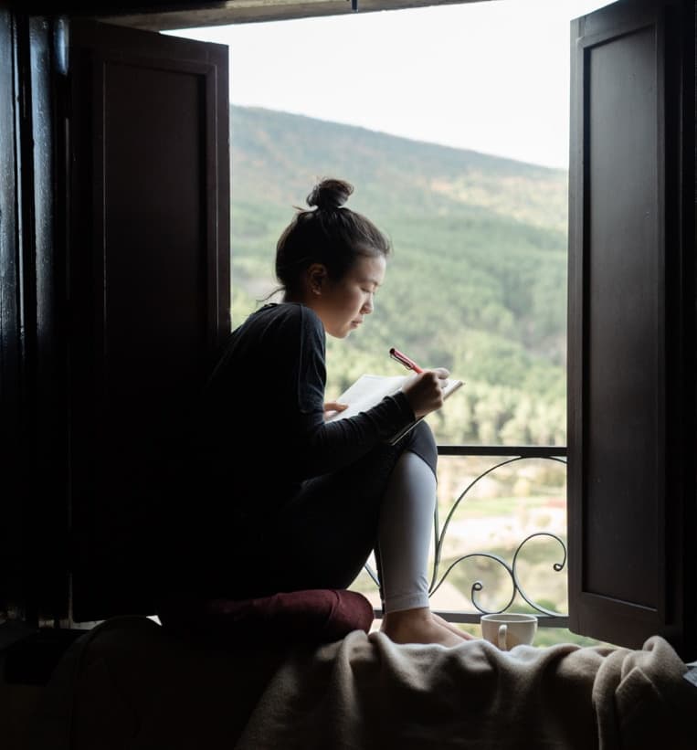 Digital nomad prioritizing her wellbeing by journaling from the window of her rural coliving space overlooking nature.