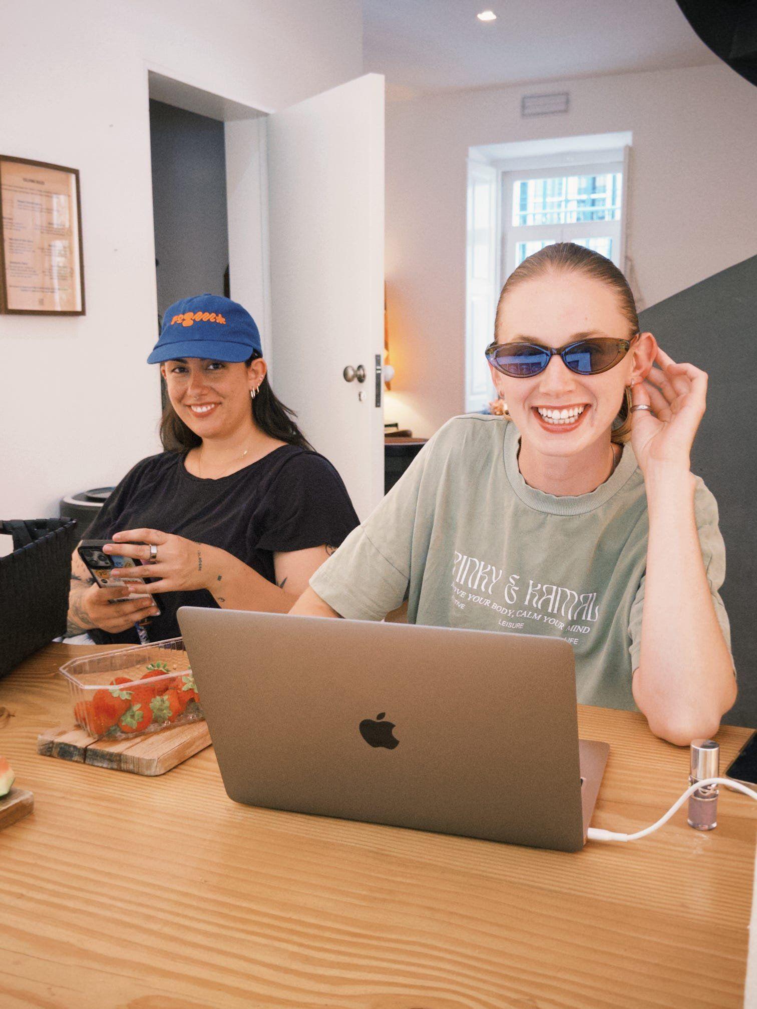 Two community members smiling at a wooden table in the coworking space, one with a laptop