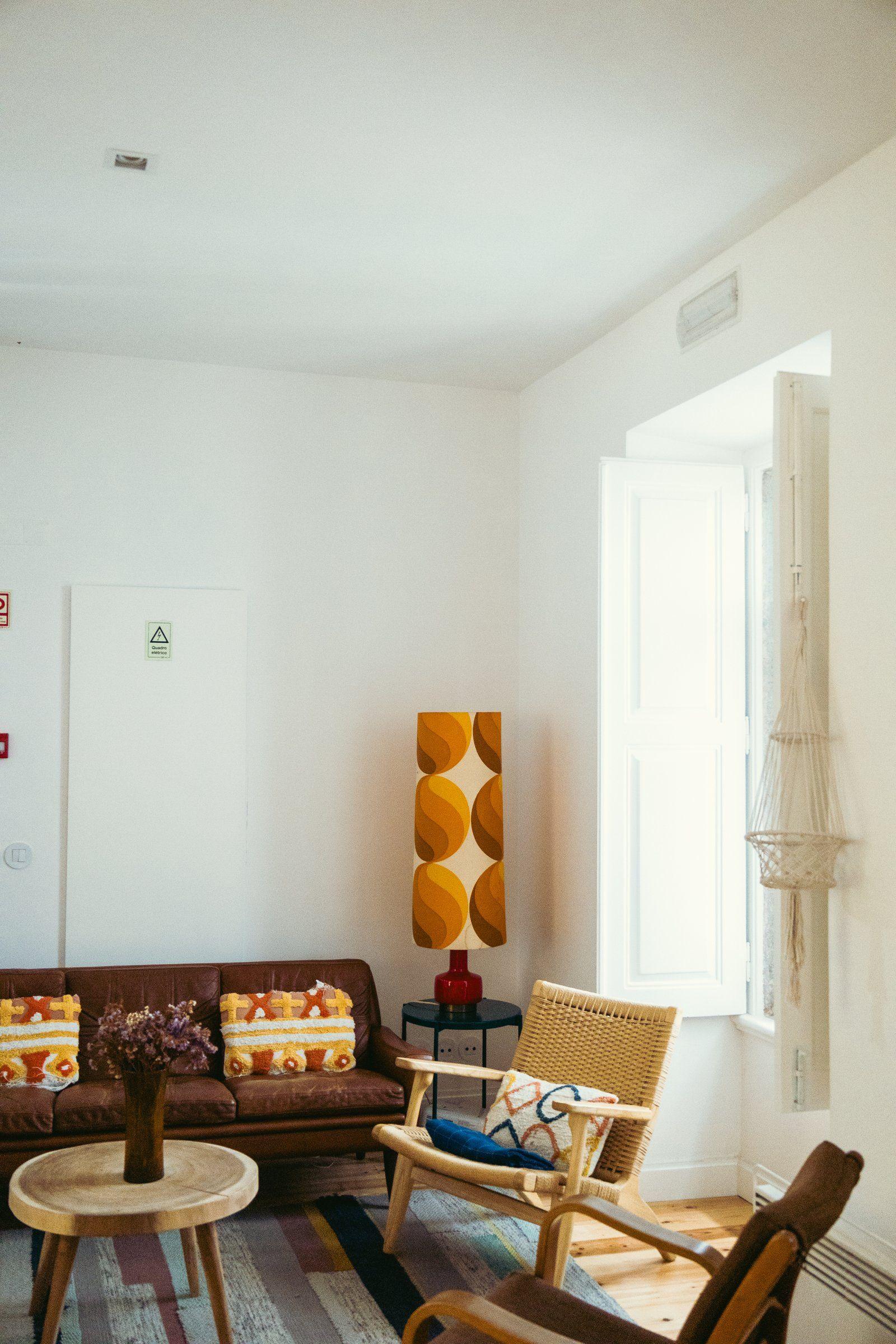 Bright shared lounge with brown leather sofa, wooden chairs, retro lamp and open window with macramé hanging