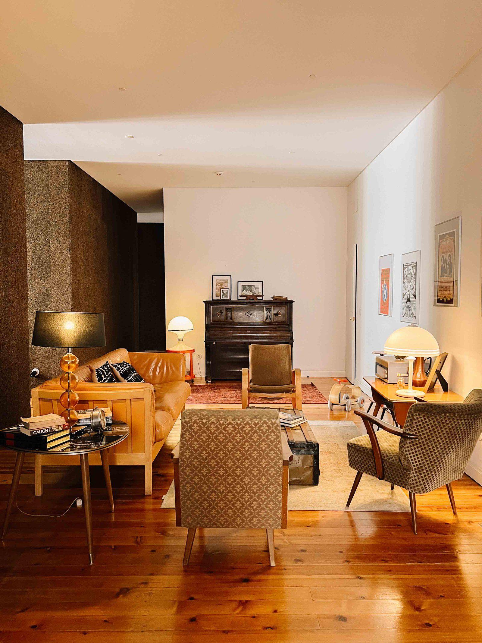Warm-lit shared living room with retro furniture, leather sofa, armchairs, wooden floor, and vintage decor including a piano