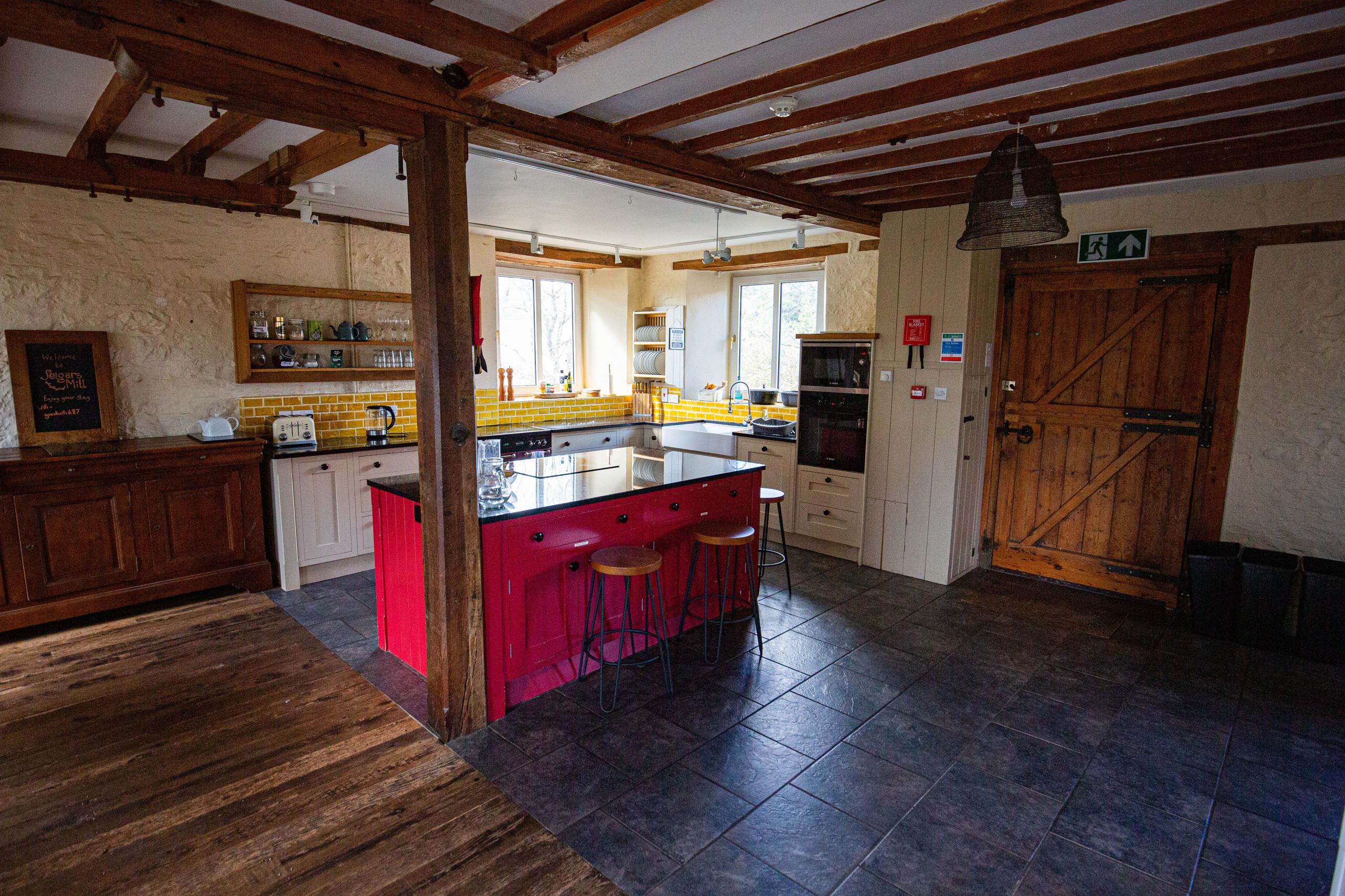 Open plan shared kitchen with red island, yellow tile backsplash, wooden beams and stone walls