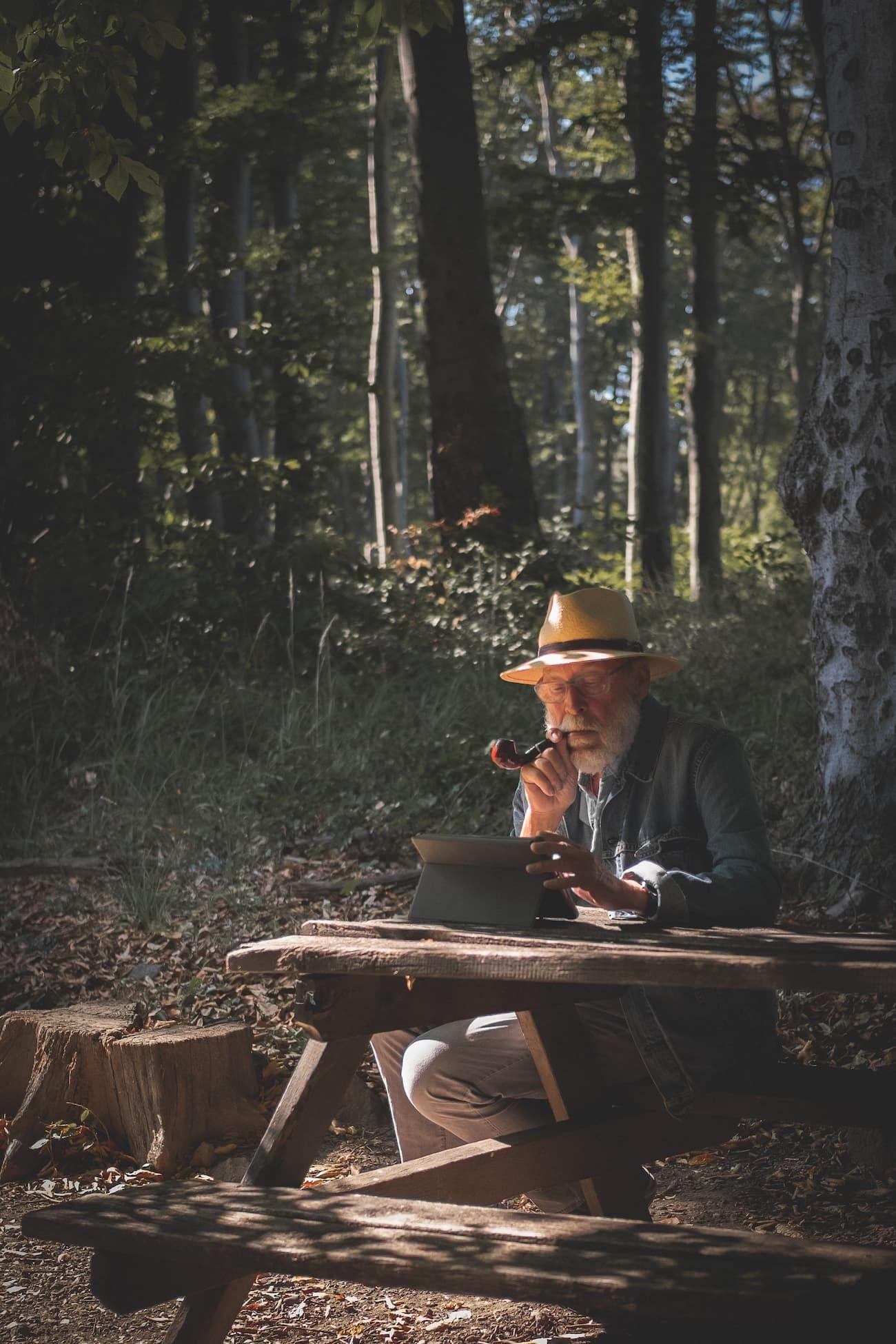 Old man with a hat and pipe working on a laptop at a wooden bench in the forest