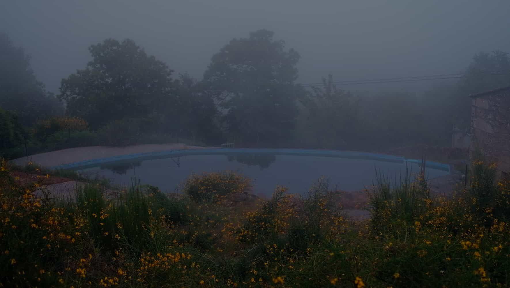 The village natural swimming pool in Senderiz on a misty morning, surrounded by yellow flowers and trees